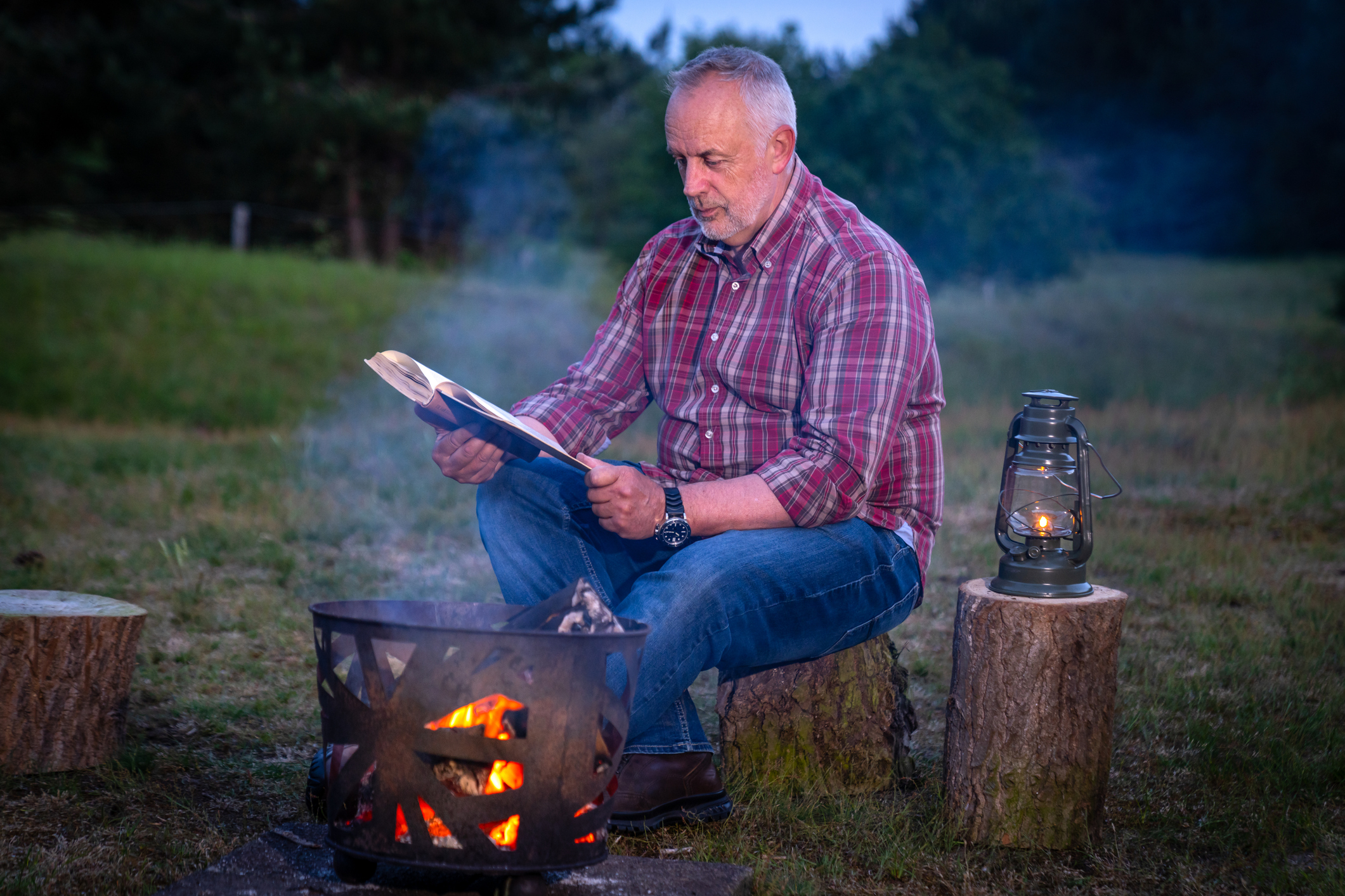 Adult male sitting outdoors on a tree trunk reading near a campfire and oil lantern