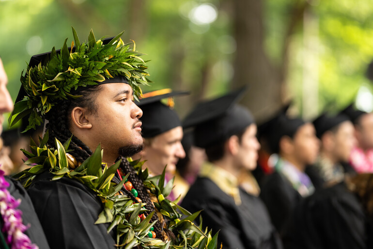 Male graduate in regalia and leis listens to commencement address.