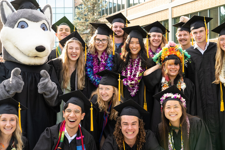 Wally the wolf mascot is surrounded by smiling graduates in regalia at Walla Walla University.