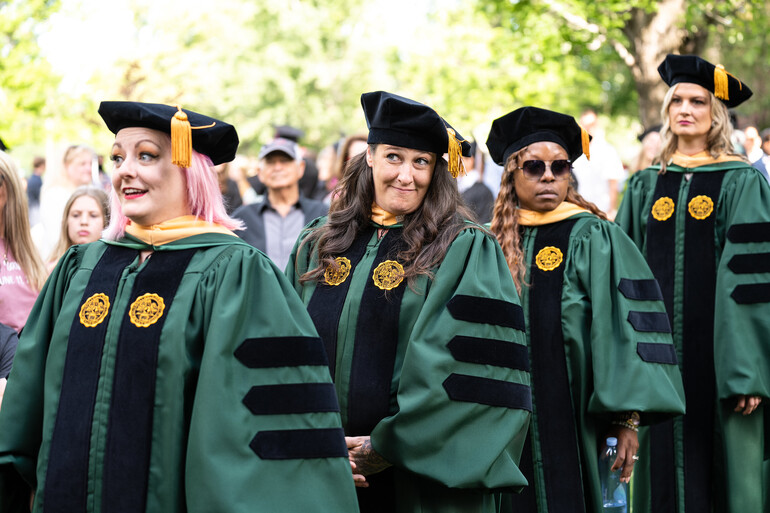 Four female doctoral graduates march in line wearing green and black regalia.