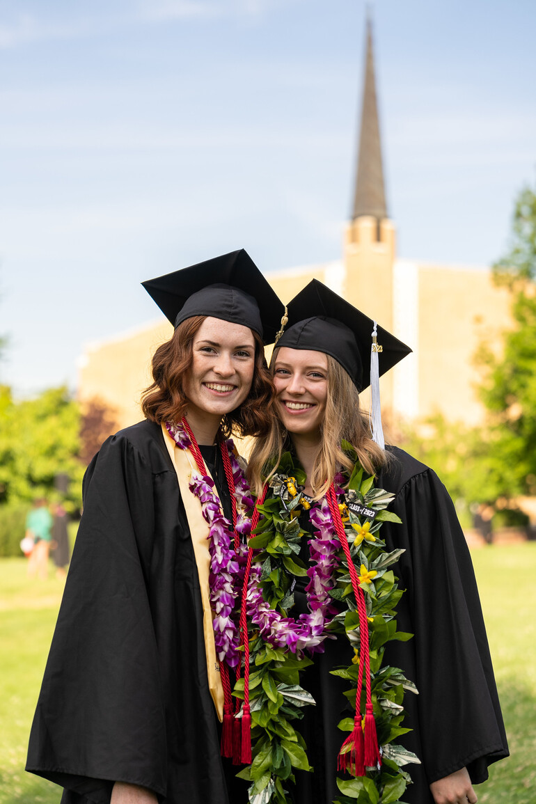 Two female graduates stand in front of church spire wearing regalia and leis.