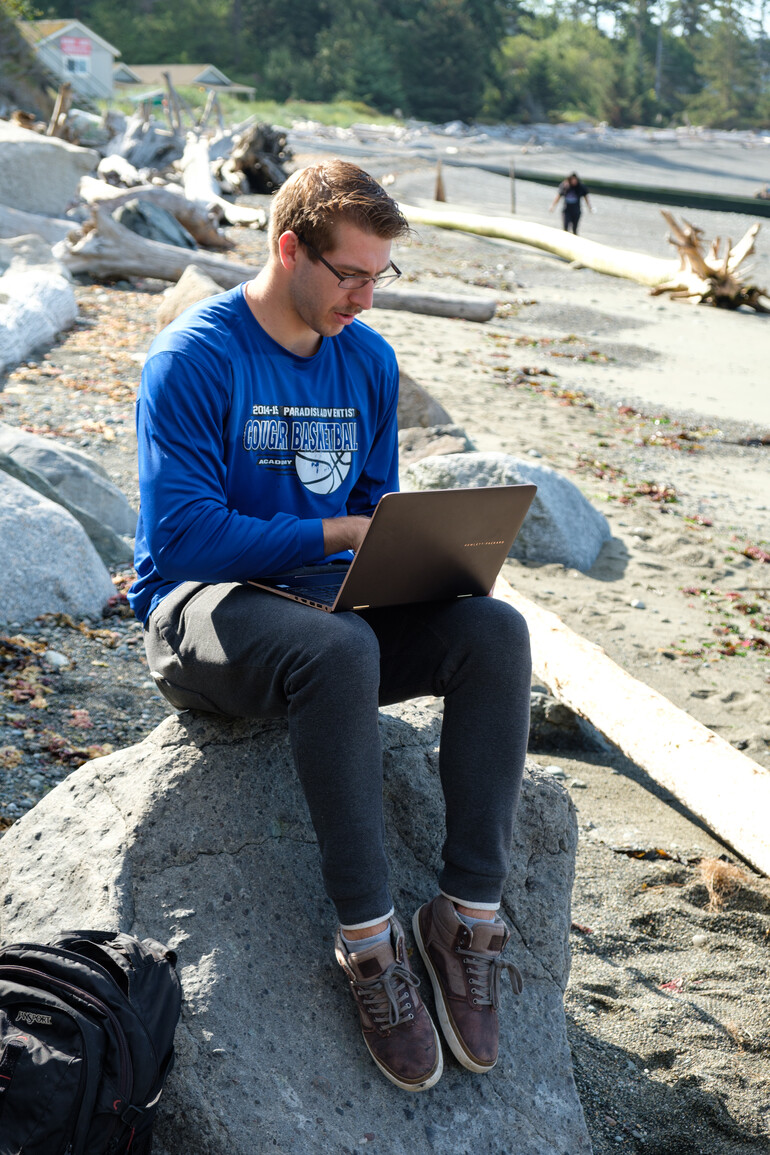 Student works on computer on sandy beach. 
