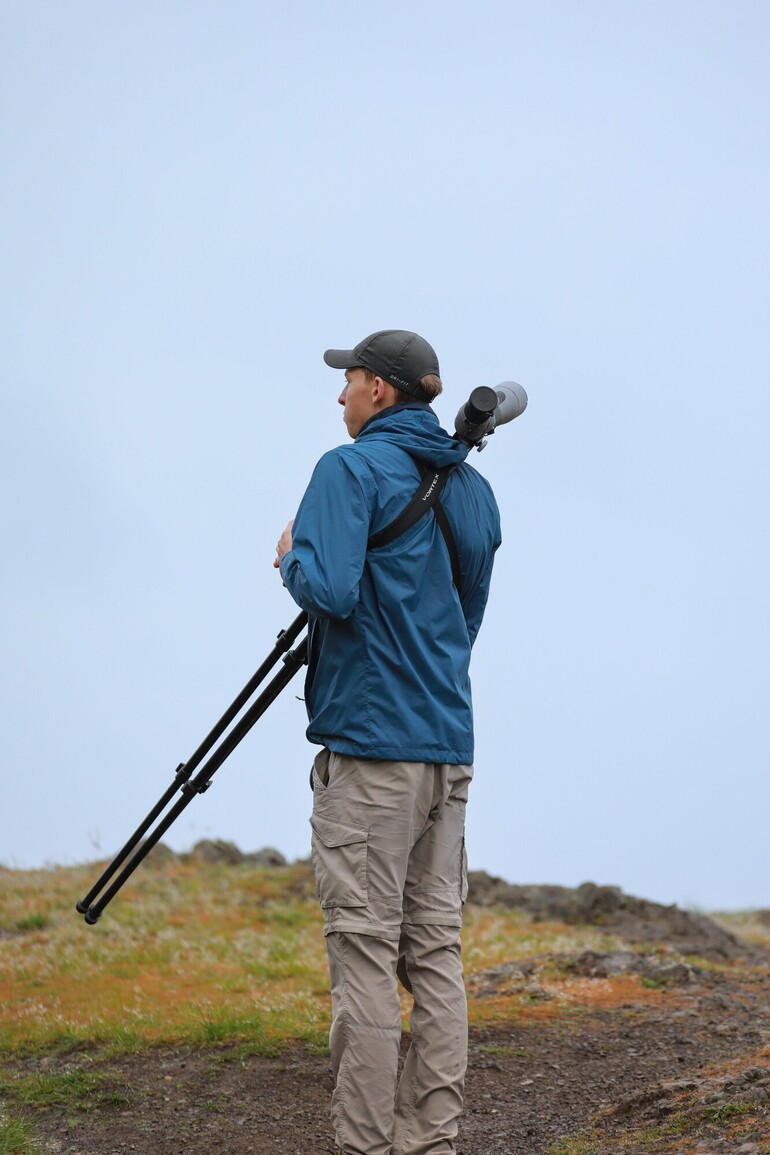 Student stands with telescope on a coastal hill.