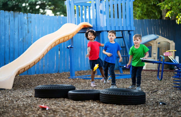 Three preschool children are having fun playing a bean bag tossing game together on the playground of their daycare