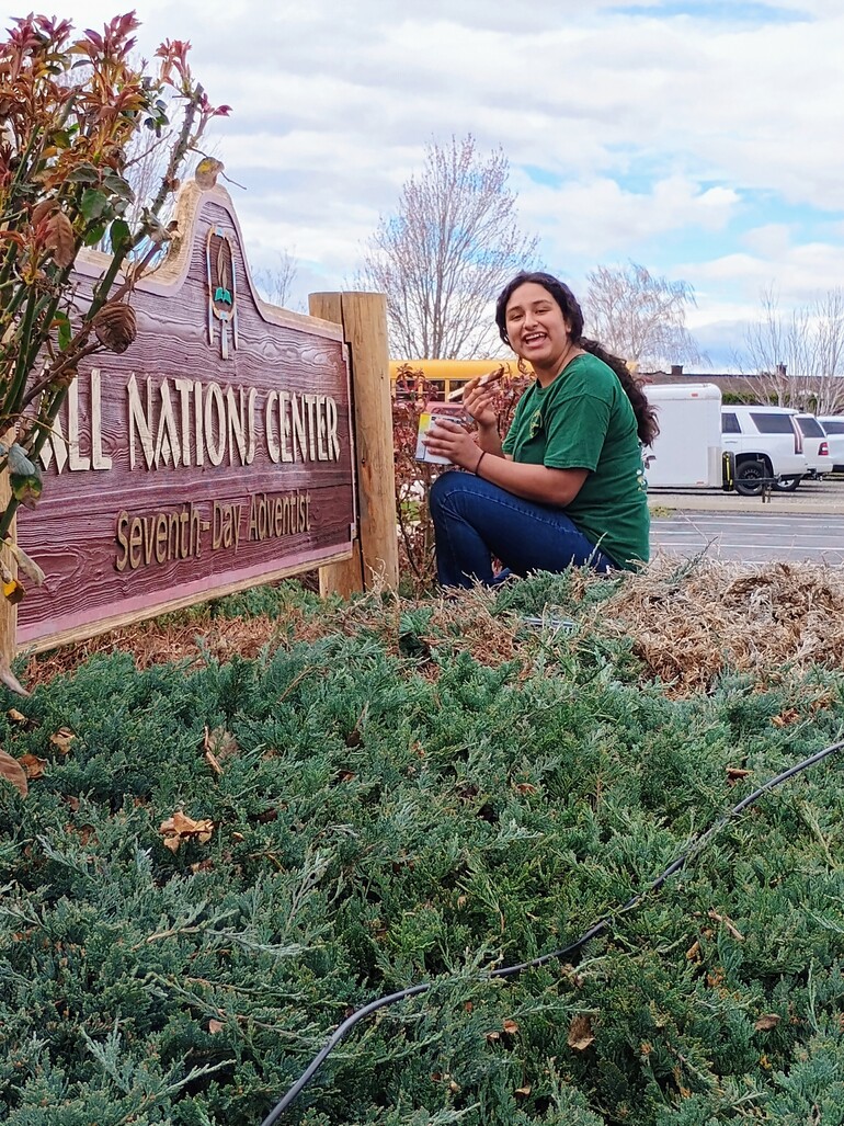 A young woman with a paint can and brush in front of a sign for the All Nations Center