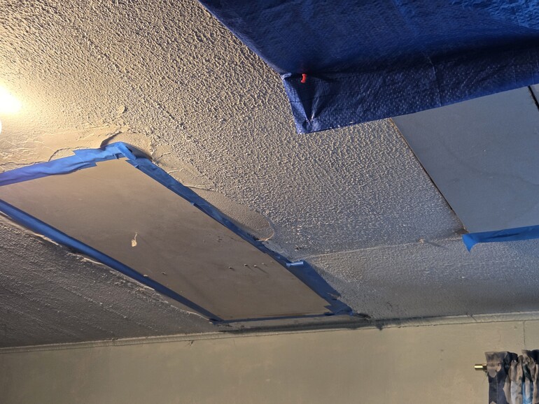 A living room ceiling with temporary repairs to water damage, including tarps, and pieces of wood secured to the ceiling.