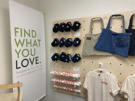 Tshirts, tote bags, posters, and chalk bags hang on wooden display next to Well-Designed Goods store sign.