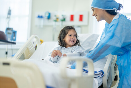 A female doctor of Indian decent checks in on a young female patient after her surgery.  The patient is sitting up in bed and appears happy.