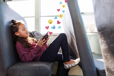 Young schoolgirl relaxes on the school bus. She is checking her social media account on her smartphone. Graphics of emojis, like buttons and heart symbols flow out from her smartphone.