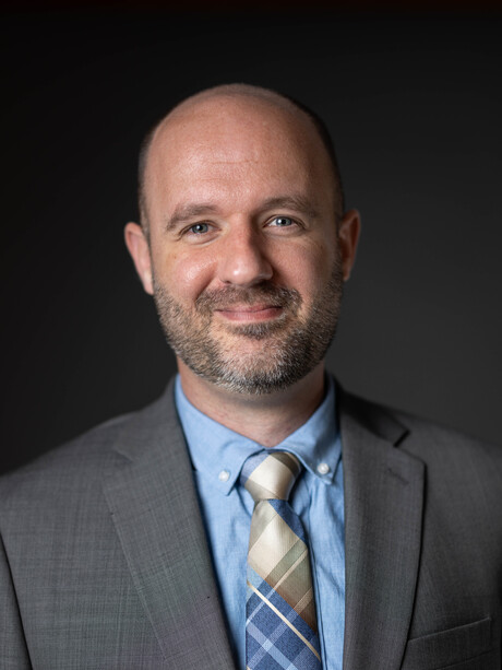 Adam Littell in a suit smiling in front of a neutral gray background