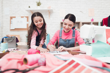 Mom and the little girl together draw a scheme of clothes. They are in the sewing workshop. They are in a good mood.