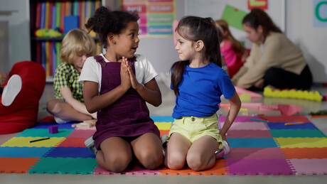 Multiracial little girls tell secrets sitting on floor in kindergarten. Adorable preschool African-American and caucasian girls sitting together in playroom share secrets