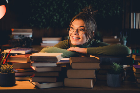 A young woman in a green jumper smiles while surrounded by books at home. The cozy indoor environment suggests a calming and enjoyable study session, perfect for exam preparation.