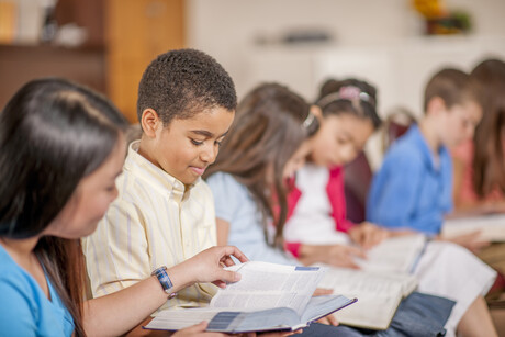 A multi-ethnic group of elementary age children are sitting together and are reading the Bible.