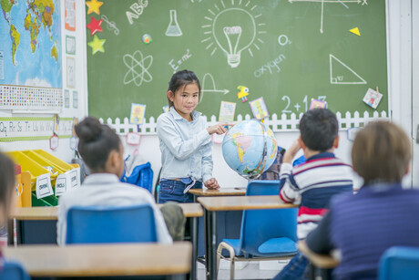 An asian elementary aged girl stands before her class and presents to them, pointing to a location on the globe.