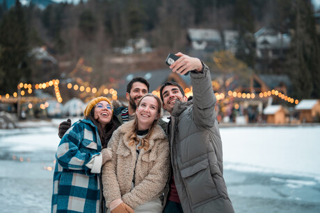 Two joyful Caucasian couples bundled in warm winter clothing take a cheerful selfie outdoors in a snowy landscape adorned with twinkling festive lights.