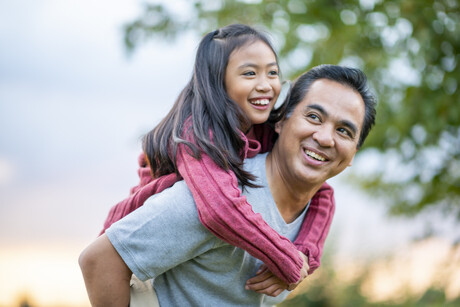 A Filipino father gives his elementary school aged daughter a piggy back ride, while they enjoy a walk outside.  They are smiling and enjoying each others company and his daughter is looking down at him.