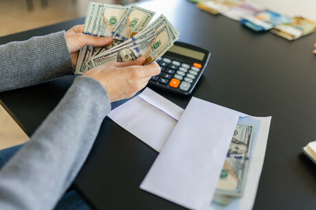 A woman counting a hundred dollar bills. There is a calculator and an envelope with some money in it next to her on the table.