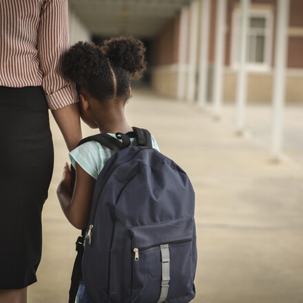 Elementary age, African American girl holds mom or teacher's hand before school begins.  She wears a backpack and clings to mom with uncertainty about starting school.