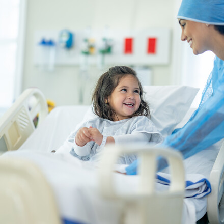 A female doctor of Indian decent checks in on a young female patient after her surgery.  The patient is sitting up in bed and appears happy.