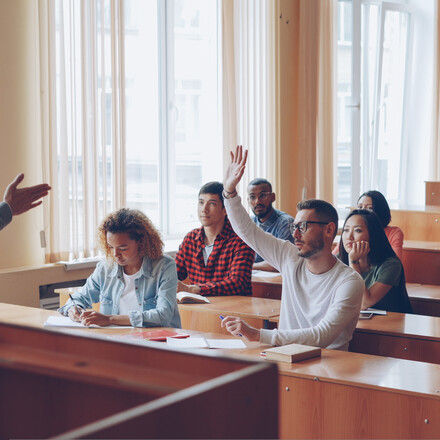 Smart guy successful student is raising hand and talking to professor while fellow students are listening to them and smiling. Pupil and teacher relations concept.