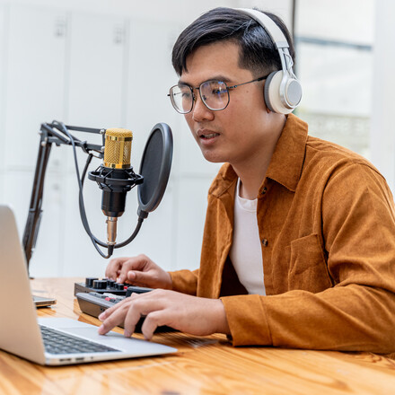 Young Asian man speaking to microphone during his live podcast while checking the feedback from his online audiences from his laptop computer