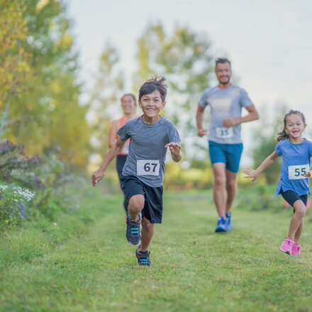 A small young mixed race family is seen participating in a race together as they run outdoors on a warm fall day.  They are each dressed comfortably in shorts and t-shirts and have race bibs on.