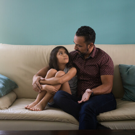 A little latin girl sitting with her father on the sofa at home and looking at him.