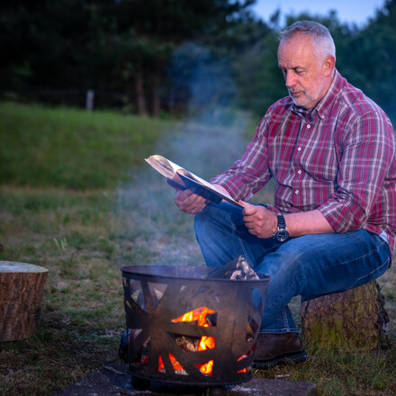 Adult male sitting outdoors on a tree trunk reading near a campfire and oil lantern