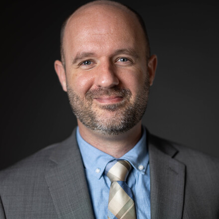 Adam Littell in a suit smiling in front of a neutral gray background