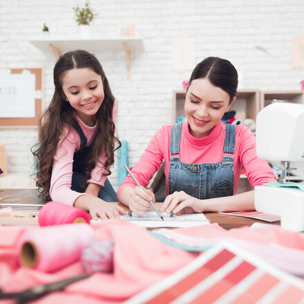 Mom and the little girl together draw a scheme of clothes. They are in the sewing workshop. They are in a good mood.
