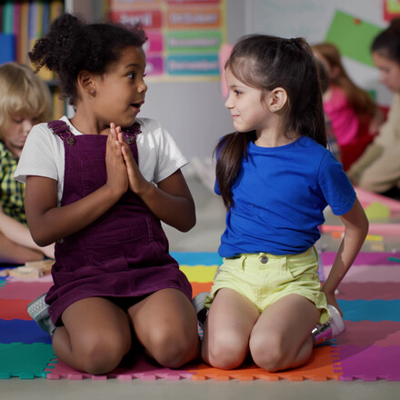 Multiracial little girls tell secrets sitting on floor in kindergarten. Adorable preschool African-American and caucasian girls sitting together in playroom share secrets