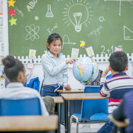An asian elementary aged girl stands before her class and presents to them, pointing to a location on the globe.