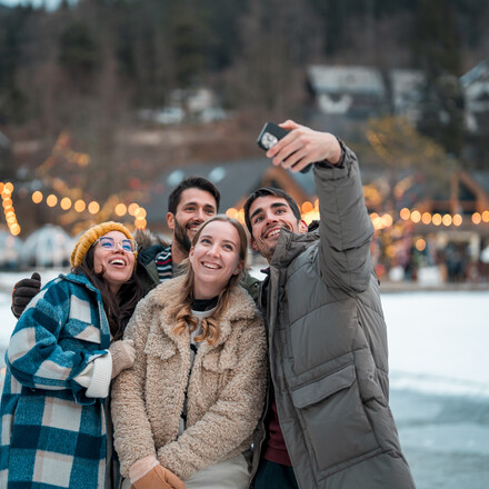 Two joyful Caucasian couples bundled in warm winter clothing take a cheerful selfie outdoors in a snowy landscape adorned with twinkling festive lights.