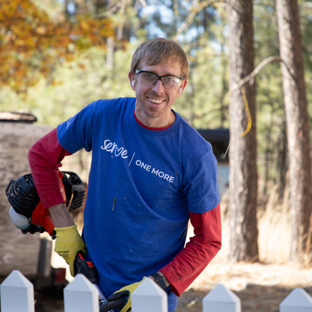 Smiling Volunteer with a Serve One More T-shirt standing in a backyard holding a String Trimmer