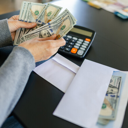 A woman counting a hundred dollar bills. There is a calculator and an envelope with some money in it next to her on the table.