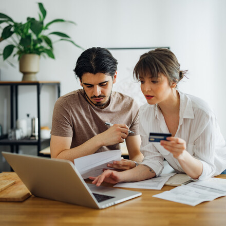 Boyfriend looking at bills while his girlfriend holding credit card and talking with him while doing home finances together online on a laptop computer in the kitchen.
