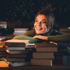 A young woman in a green jumper smiles while surrounded by books at home. The cozy indoor environment suggests a calming and enjoyable study session, perfect for exam preparation.