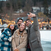 Two joyful Caucasian couples bundled in warm winter clothing take a cheerful selfie outdoors in a snowy landscape adorned with twinkling festive lights.