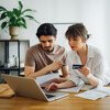 Boyfriend looking at bills while his girlfriend holding credit card and talking with him while doing home finances together online on a laptop computer in the kitchen.