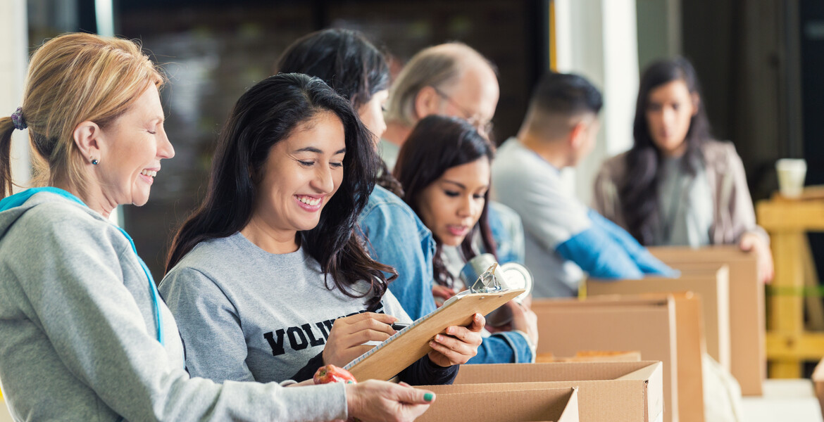 Mature adult Caucasian woman is standing with mid adult Hispanic woman as they pack cardboard boxees full of donated food in charity food bank. Other volunteers are lined up behind them, also sorting donated groceries into boxes. Hispanic woman is writing on checklist on clipboard.