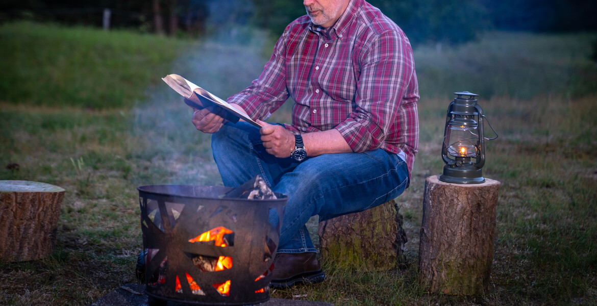 Adult male sitting outdoors on a tree trunk reading near a campfire and oil lantern