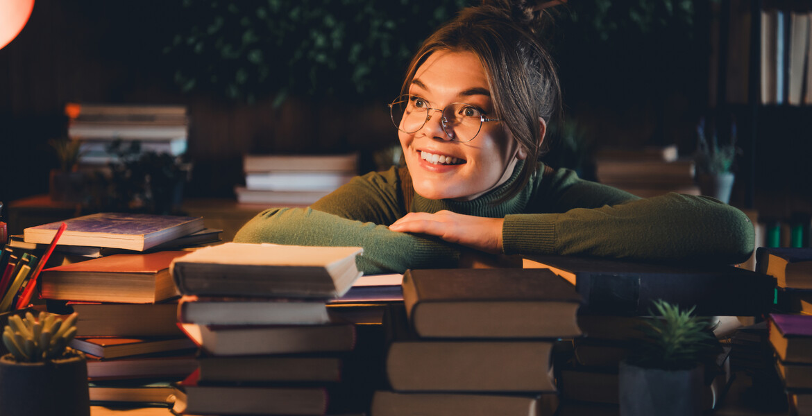 A young woman in a green jumper smiles while surrounded by books at home. The cozy indoor environment suggests a calming and enjoyable study session, perfect for exam preparation.
