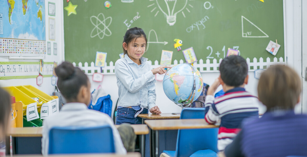 An asian elementary aged girl stands before her class and presents to them, pointing to a location on the globe.