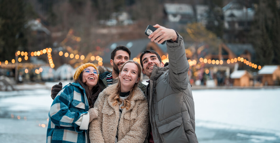 Two joyful Caucasian couples bundled in warm winter clothing take a cheerful selfie outdoors in a snowy landscape adorned with twinkling festive lights.