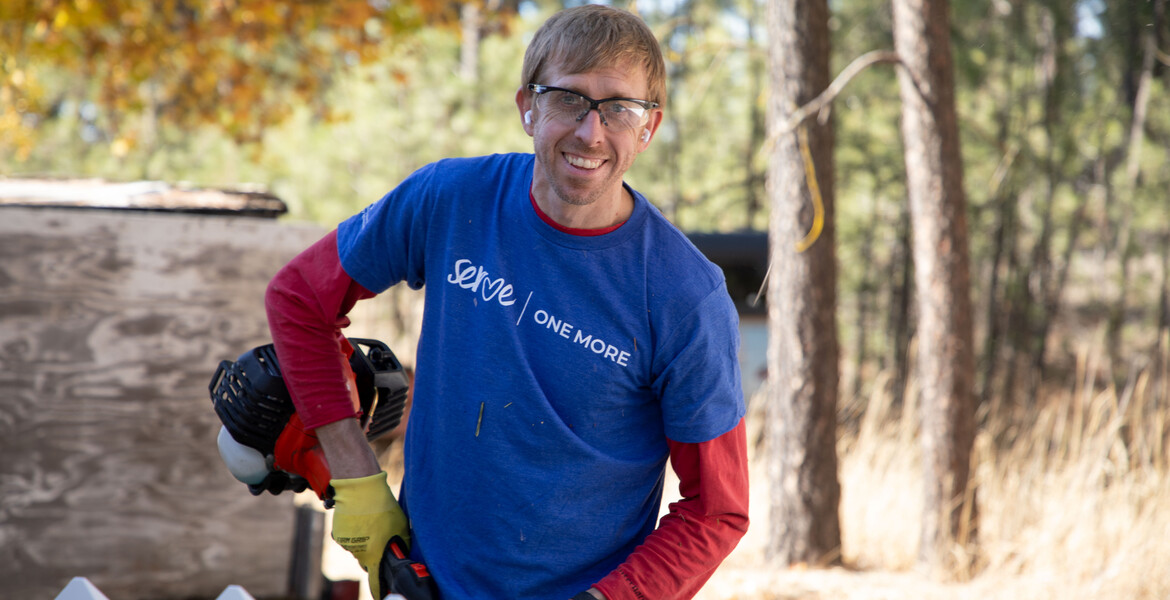 Smiling Volunteer with a Serve One More T-shirt standing in a backyard holding a String Trimmer