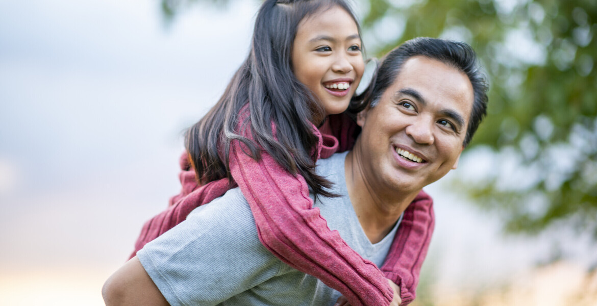 A Filipino father gives his elementary school aged daughter a piggy back ride, while they enjoy a walk outside.  They are smiling and enjoying each others company and his daughter is looking down at him.