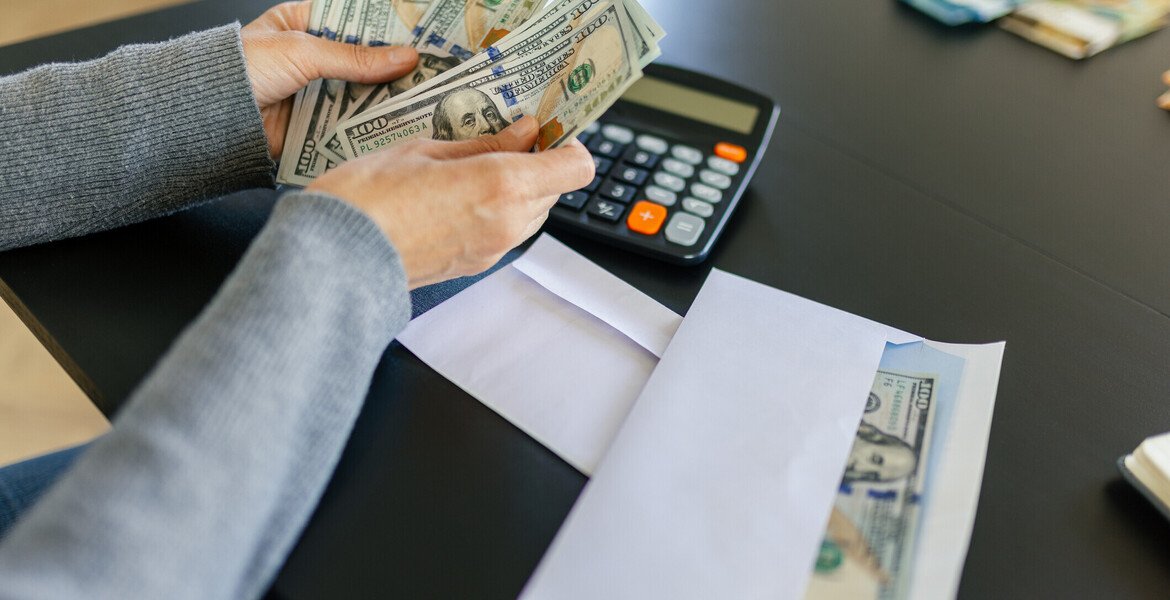 A woman counting a hundred dollar bills. There is a calculator and an envelope with some money in it next to her on the table.