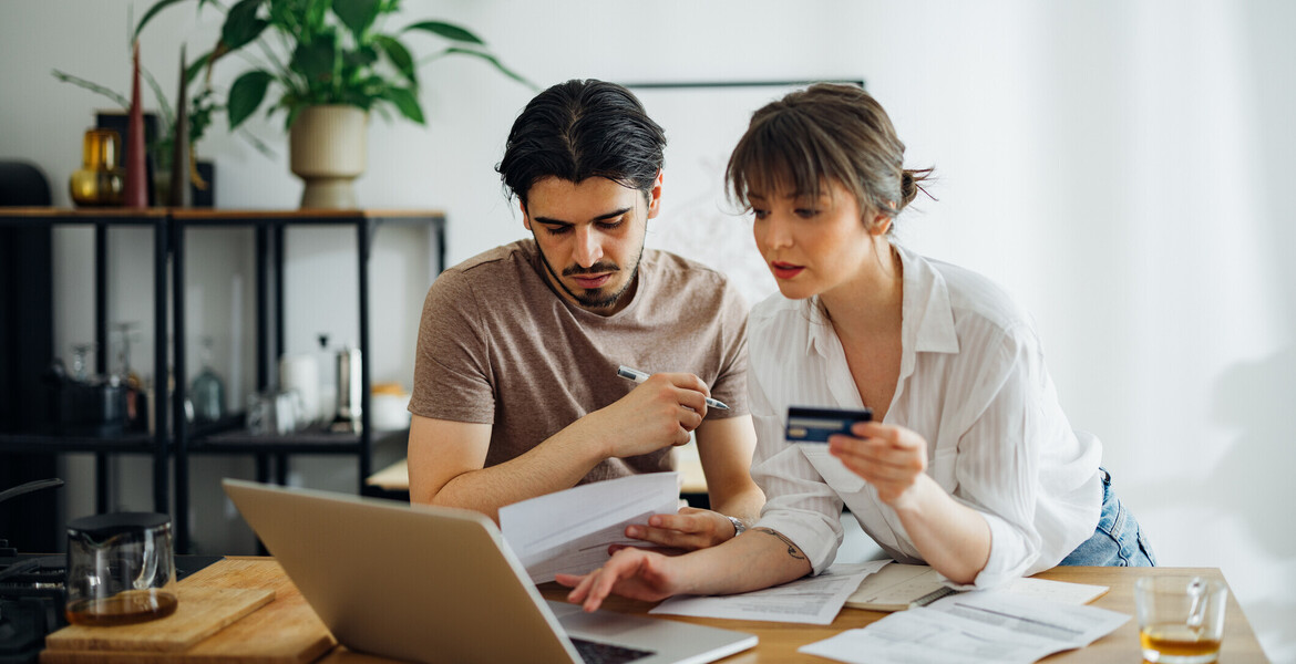 Boyfriend looking at bills while his girlfriend holding credit card and talking with him while doing home finances together online on a laptop computer in the kitchen.